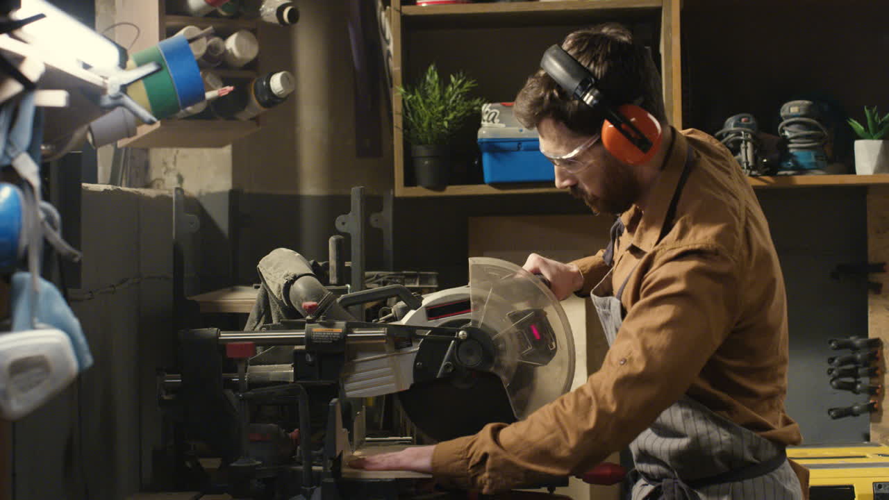 joven carpintero caucásico con gafas y auriculares cortando tablones de madera con una sierra circular eléctrica en un taller de carpintería