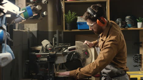 young caucasian male carpenter in goggles and headphones cutting wooden plank with a circular electric saw in carpentry workshop