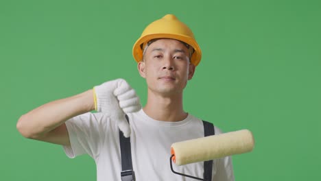 close up of asian man painter wearing safety helmet showing thumbs down gesture to camera while standing in the green screen background studio
