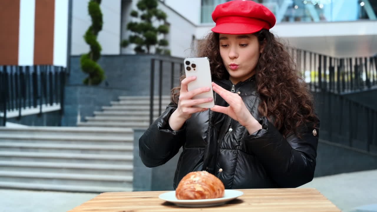 Una giovane blogger influencer con un cappello rosso che fotografa un croissant con il suo smartphone. Rallentatore