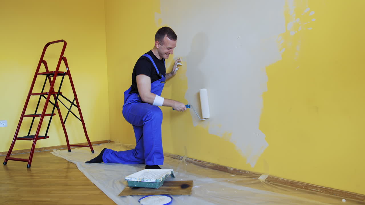 Happy male worker is painting wall. Professional young man with paint roller having fun in the apartment. Room painting job. Renovation home concept.