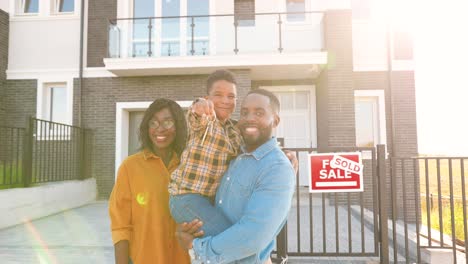 retrato de una familia afroamericana feliz con un niño pequeño parado en una casa nueva en un suburbio y mostrando las llaves de la cámara