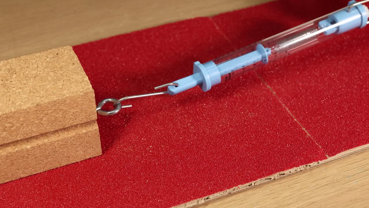 A hand uses a spring scale to measure friction on a wooden block over sandpaper, demonstrating force dynamics