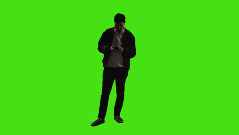 hombre con auriculares enviando o leyendo mensajes de texto o desplazándose en línea en el teléfono móvil de pie contra la pantalla verde con iluminación de bajo nivel