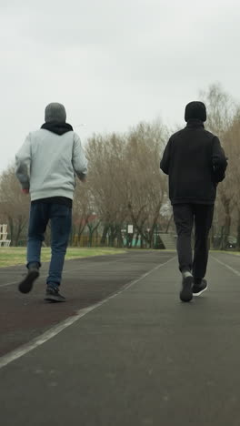 back view of an adult and two young boys jogging side by side on a track in a stadium, with a residential building visible across the road, with bare trees on the right hand side