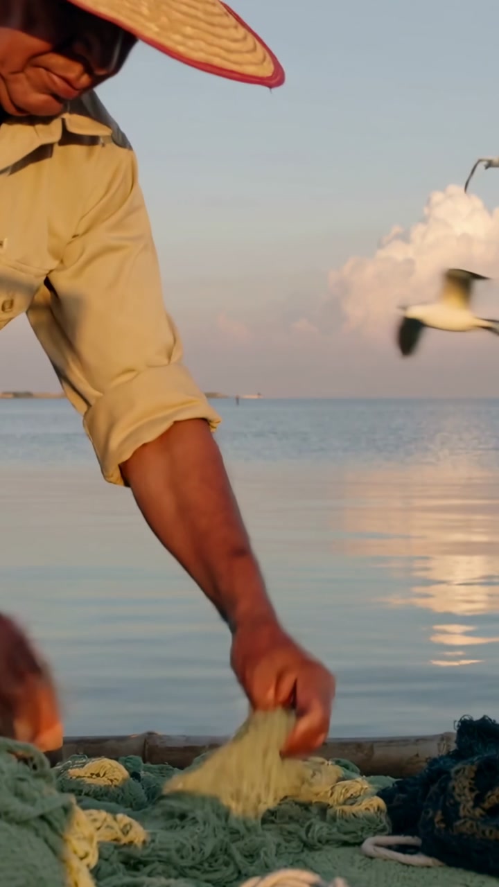 Vertical video: Leaning man in straw-hat pulling dark nets on boat at sea, sorting nets, copy space