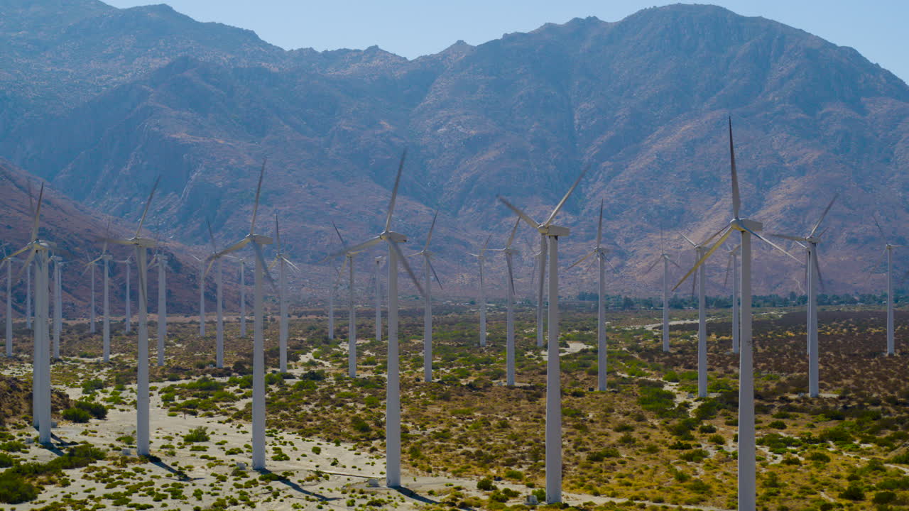 Palm Springs windmills spinning with the rugged San Jacinto Mountains rising behind them in California under a clear blue sky on a bright, sunny day. A vivid, modern-meets-nature scene