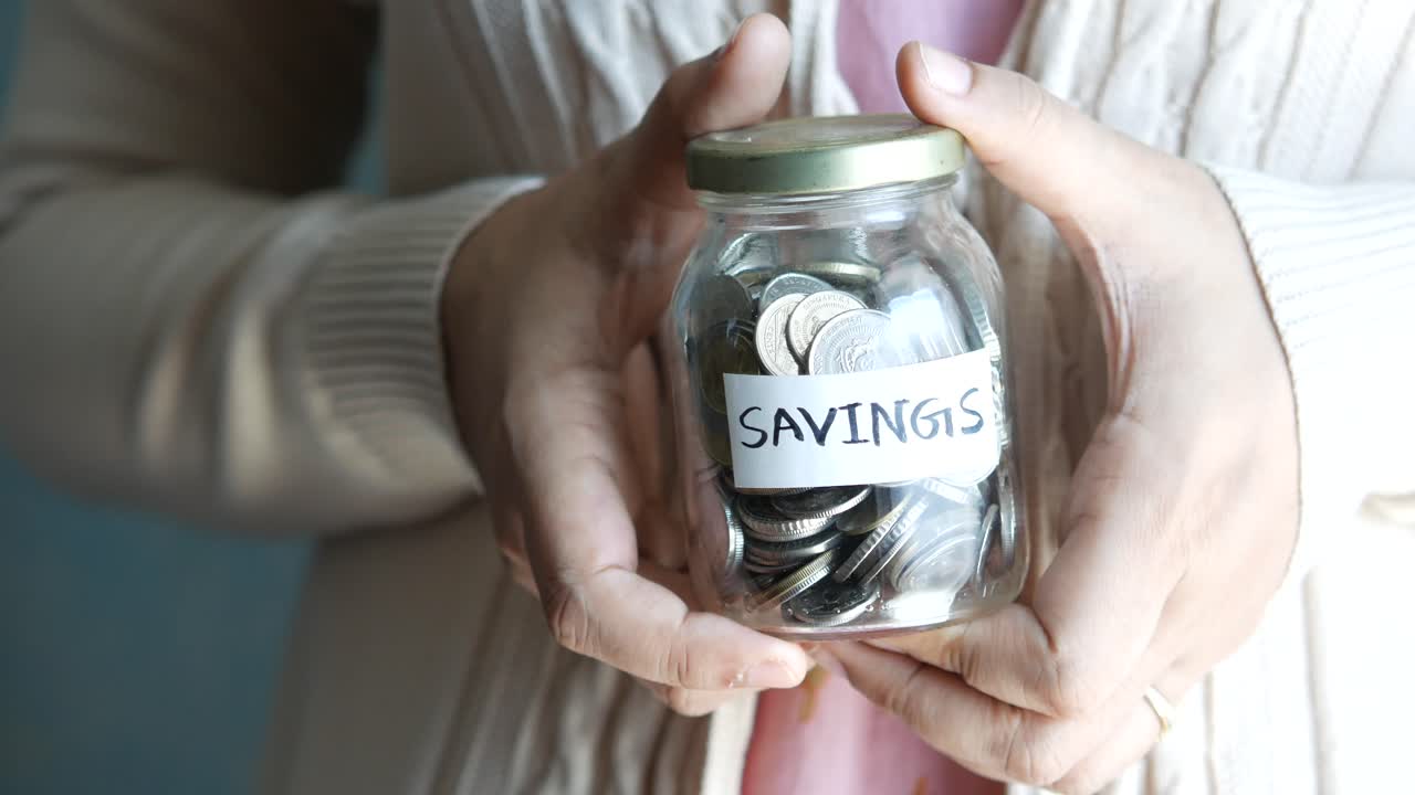 Person holding a glass jar with coins and the word "SAVINGS" written on it.