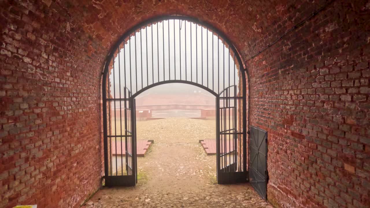 Besøkende går gjennom en gammel mursteinsbuegang med jerngrinder. Utsikten avslører et tåkete landskap bortenfor. Atmosfæren fanger essensen av historie og natur.