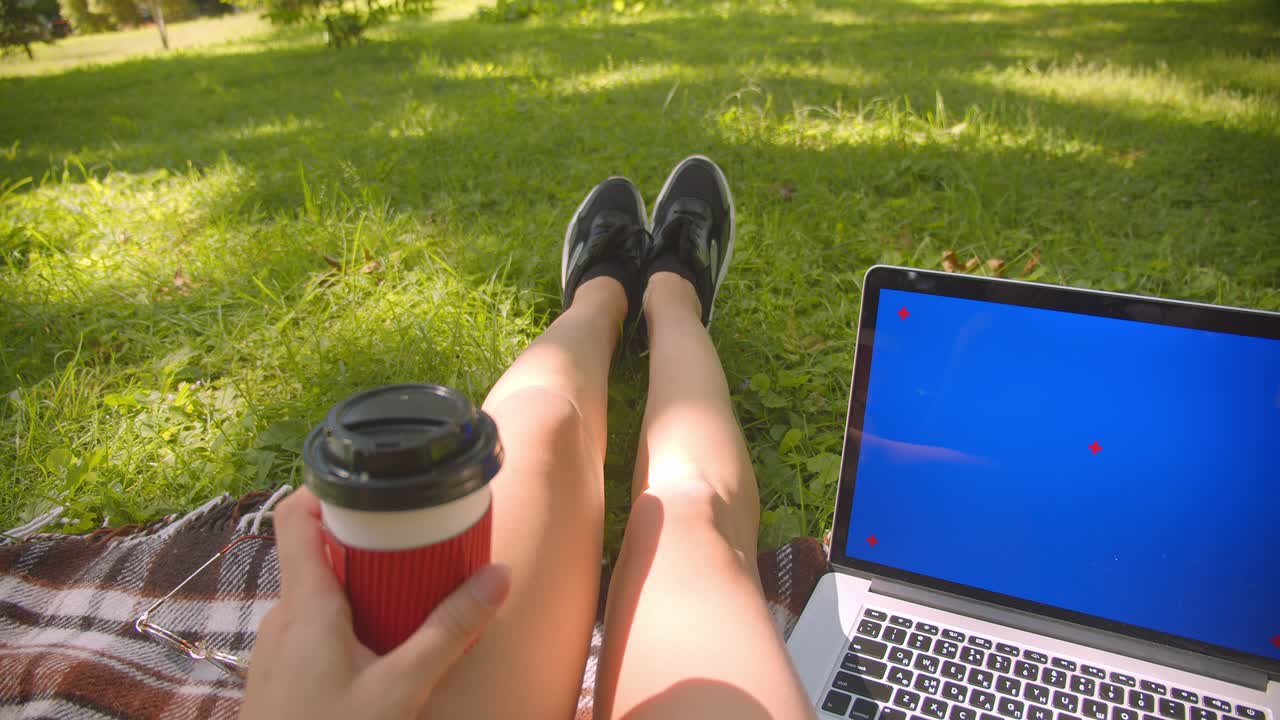 retrato en primer plano de una joven con piernas caucásicas bonitas sentada en una alfombra sosteniendo un café usando una computadora portátil con pantalla azul en el parque al aire libre