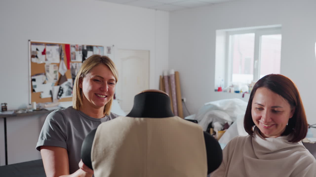 Creative minds shake hands while tweaking beige waistcoat on black mannequin, sharing smiles inside light filled sewing studio lined with fabric rolls, mood board, scattered equipment