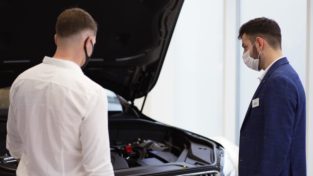 consultor que muestra el motor del coche bajo el capó abierto a un cliente en la sala de exposiciones de automóviles durante el bloqueo, vista trasera