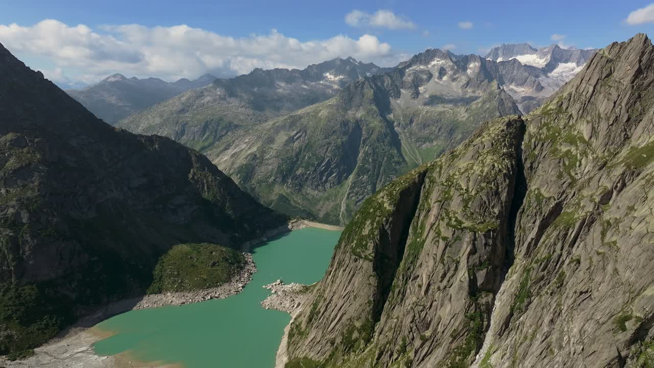 Alpine reservoir with turquoise water, lies between steep rocky cliffs, held back by a dam, towering mountain peaks rise in the distance, aerial shot