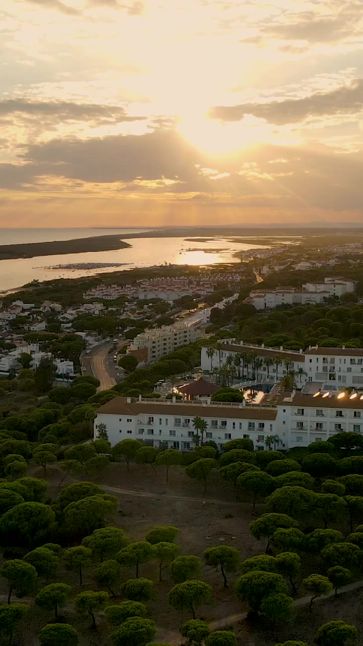 Betagende luftoptagelser af El Rompido, Spanien, i den gyldne time, der fremviser kystbyen, floden og frodige fyrretræer under en smuk solnedgang.