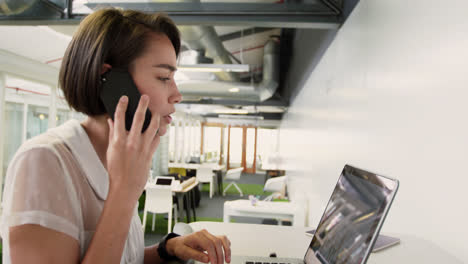 Businesswoman-talking-on-mobile-phone-while-using-laptop-in-modern-offi-4k