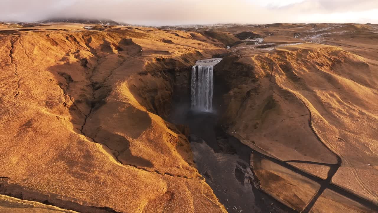 Aerial view of Iceland's Skógafoss waterfall cascading through a rugged, golden landscape. The Skógá River carves its path. Perfect for travel, nature documentaries, and scenic visuals.
