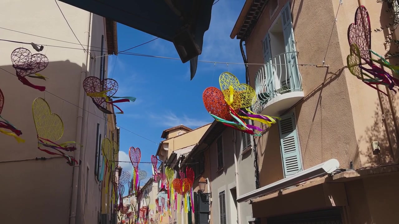 Cuori intrecciati luminosi e nastri fluttuanti pendono sopra una strada stretta fiancheggiata da edifici color pastello, balconi e persiane sotto un cielo azzurro vivido