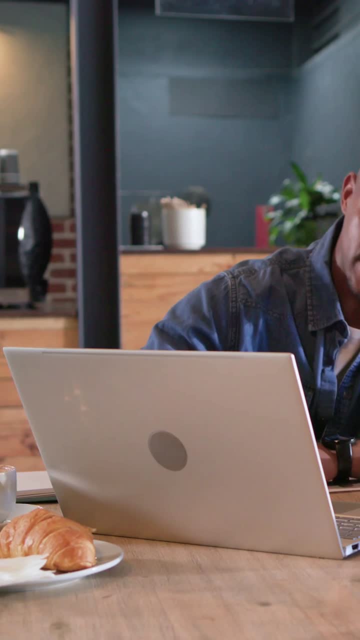 Vertical video: Picking papers man in denim shirt flipping pages and working on silver laptop at c