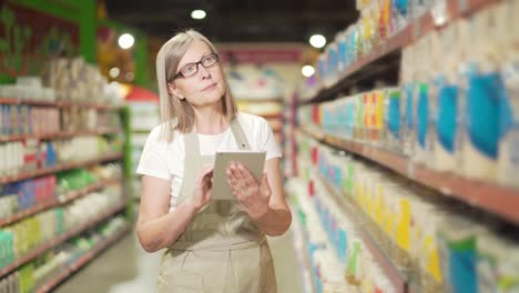 empleada gerente sénior en un supermercado, tienda de comestibles o delicatessen con un delantal con una tableta digital. empleada madura propietaria de una pequeña empresa inspecciona los estantes de productos de inventario haciendo el inventario de comprobación