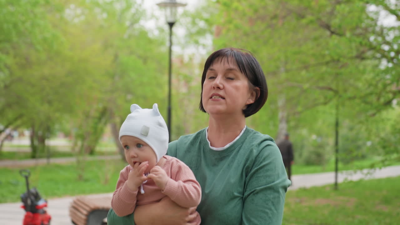 Elderly Woman Sings To Child Outdoors, Senior Woman Gently Sings To Baby In Spring Park, Elderly Grandmother Tenderly Sings Lullaby To Her Baby Amid Lush Park Landscape With Birch Woods