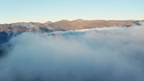 Mountains-peaking-through-a-blanket-of-clouds-at-sunrise,-aerial-view