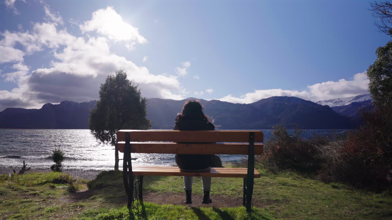 Young girl sits silently on wooden bench facing vast lake, surrounded by mountain silhouettes and scattered clouds