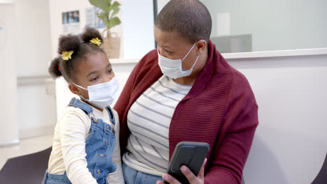 Madre-E-Hija-Afroamericanas-Usando-Máscaras-Faciales-Usando-Un-Teléfono-Inteligente-En-El-Hospital,-En-Cámara-Lenta