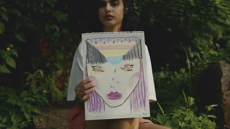 young woman sits with a drawn self-portrait on a stone in nature