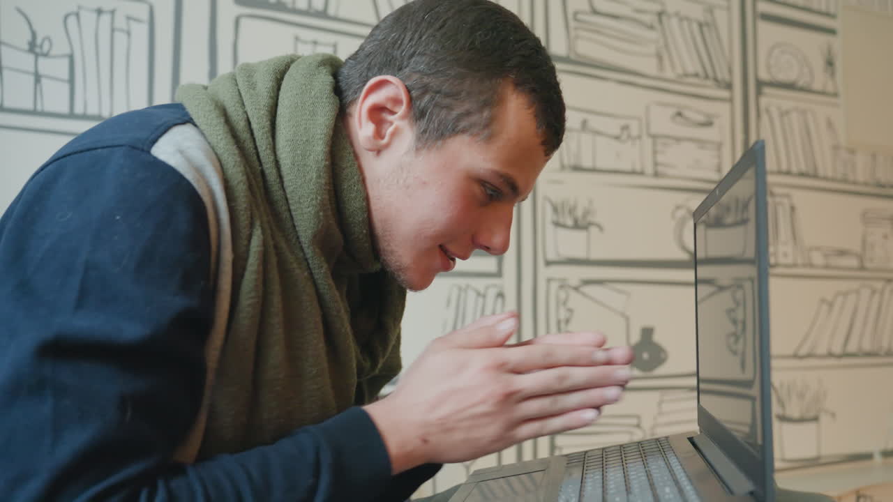 Uomo bianco che si china verso il laptop, espressione curiosa e sorriso dolce, sciarpa verde e maglione casual, sfondo con carta da parati a libreria illustrata, primo piano delle mani che digitano sulla tastiera.