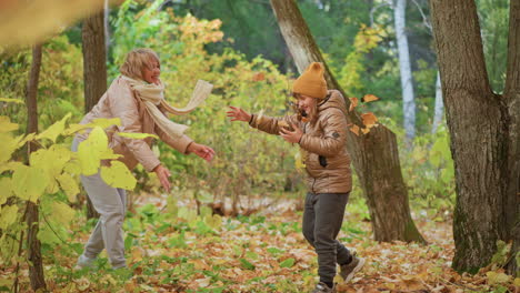 joyful mother tosses dry autumn leaves playfully onto her laughing daughter during lighthearted moment in forest filled with golden foliage, both wearing warm clothes on beautiful sunny fall day