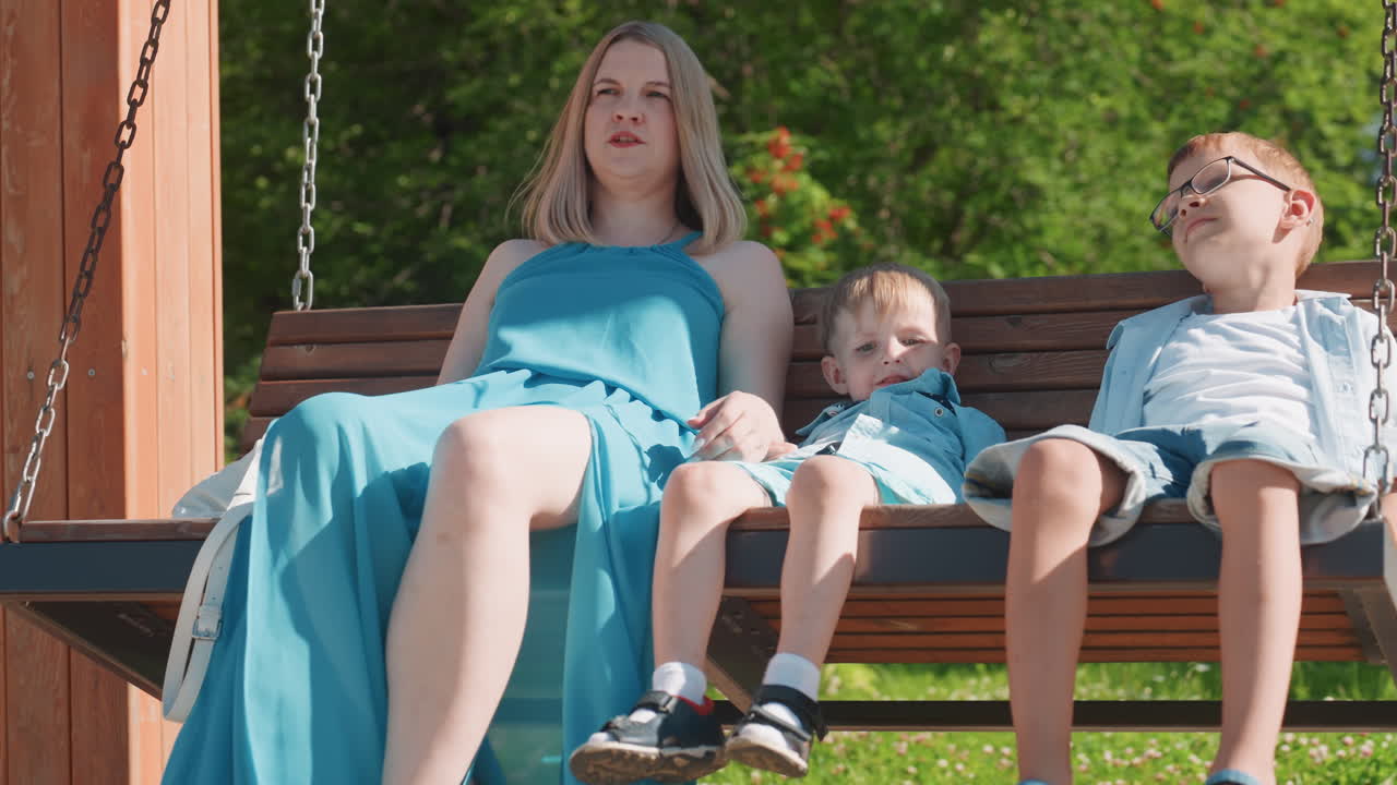 Fredfyldt scene med mor og børn udendørs. Mor og børn sidder fredeligt på en gynge i parken. Et roligt øjeblik, hvor mor og børn nyder en blid udendørs fritidsaktivitet.