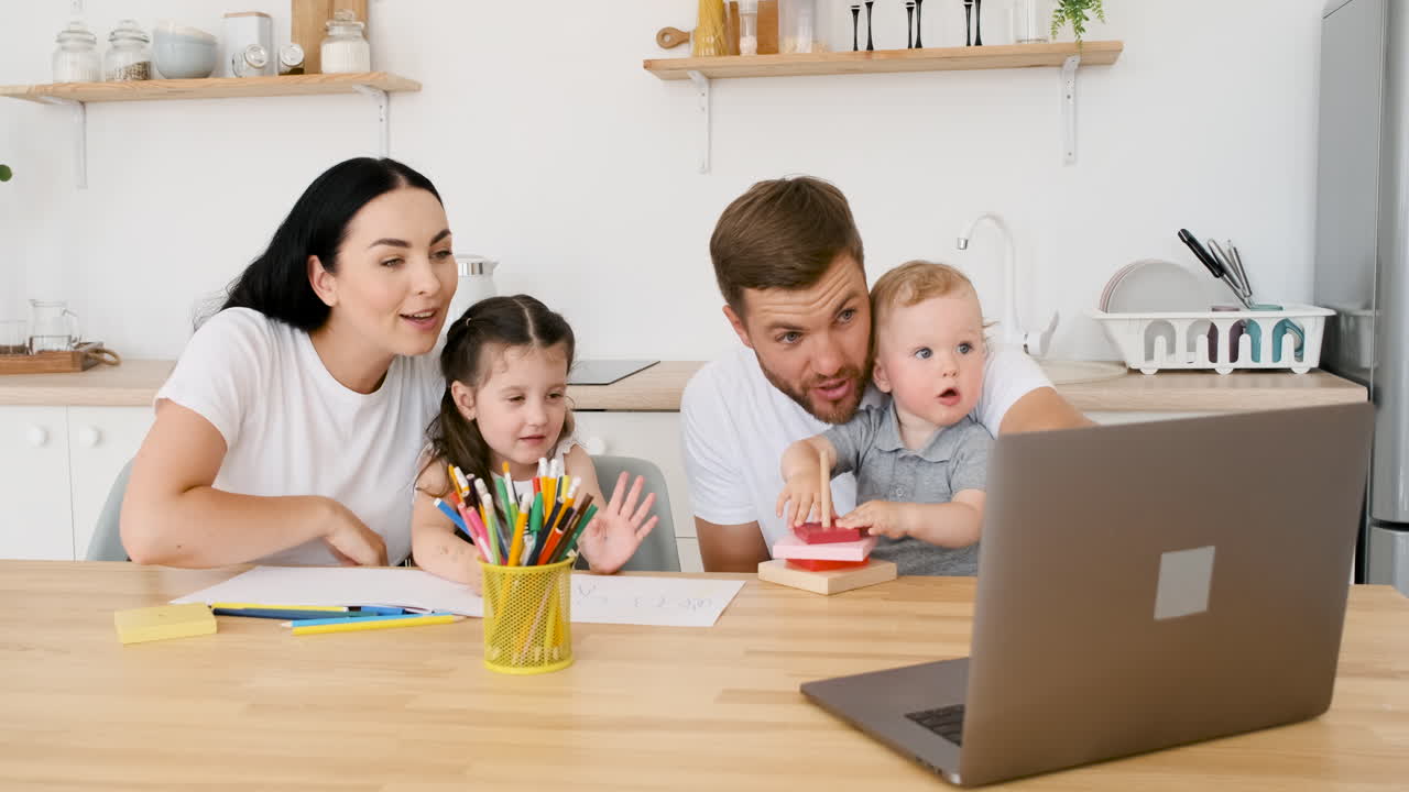 현대적인 노트북으로 화상 통화를 하면서 두 자녀와 놀고 있는 행복한 부모가 부엌 테이블에 앉아 있습니다.