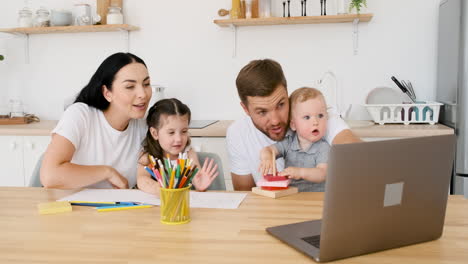 padres felices sentados en la mesa de la cocina jugando con sus dos hijos mientras hacen una videollamada en una laptop moderna