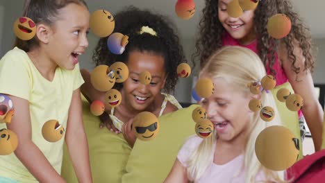 animation of various emojis moving over happy diverse schoolgirls using tablet together at beak time