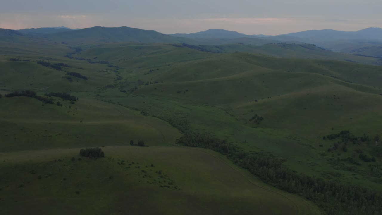 Filmato drone di vaste colline verdi ondulate, valli tortuose e creste montuose distanti sotto la morbida luce della sera nella Repubblica dell'Altai, Russia, ideale per temi di viaggio e natura.