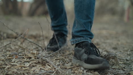 close-up shot of a person walking on a forest path, wearing black sneakers and blue jeans, the ground is covered with leaves and twigs