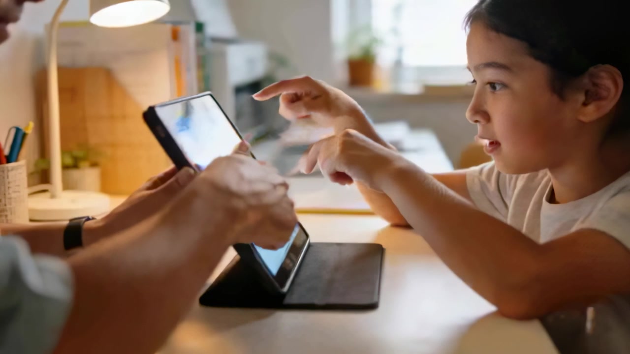 Pointing child tapping tablet at home desk while adult steadying folio case, guiding learning