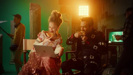 beautiful smiling actress wearing renaissance dress and actor wearing motion capture suit having lunch break, sitting on chairs, use laptop and talk. studio high budget movie. costume drama film set