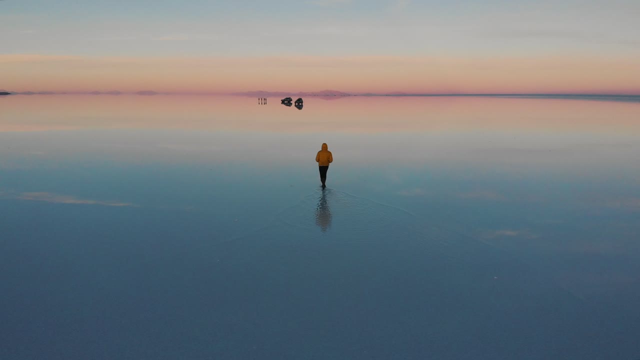 A young man with a yellow jacket walking on flooded salt flat in bolivia during sunrise