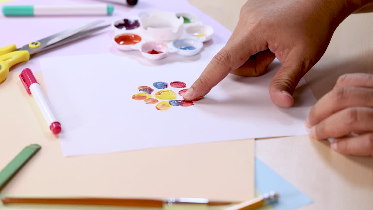 Finger dabs paint, creates colorful fingerprints on paper, surrounded by art tools under bright lighting