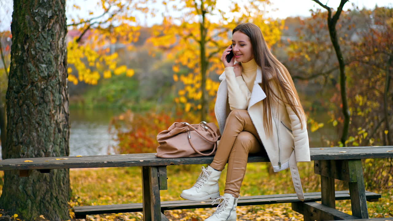 Calm relaxed lady in white jacket and beige leggings speaks on her phone. Resting woman spending time in the autumn park sitting on the wooden table.