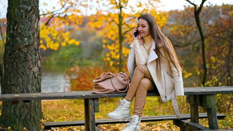 calm relaxed lady in white jacket and beige leggings speaks on her phone. resting woman spending time in the autumn park sitting on the wooden table.