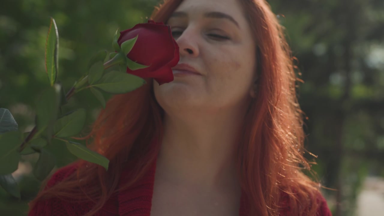 Donna con un maglione rosso scarta e tiene in mano una rosa artificiale a stelo singolo all'aperto su una pietra. La scena presenta una luce naturale soffusa, uno sfondo di fogliame verde e un primo piano sui dettagli della texture del fiore.