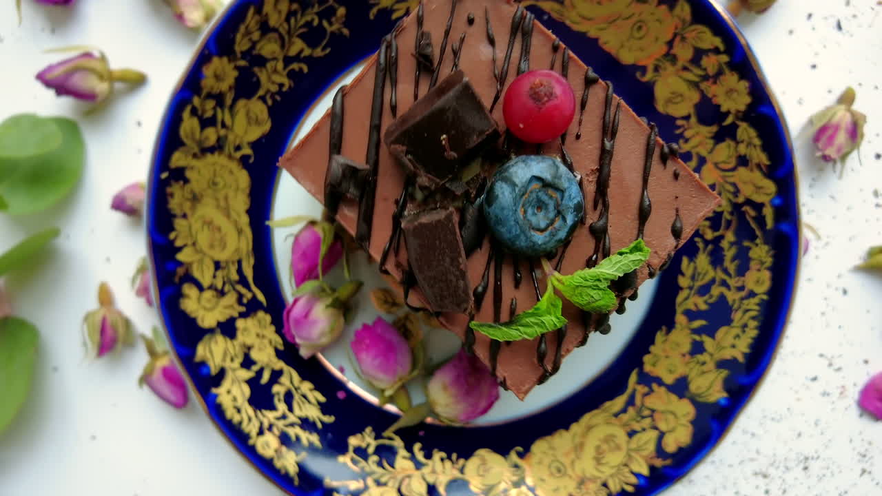 A tempting chocolate cake slice topped with blueberries, raspberries, and mint sits beautifully on an ornate blue plate surrounded by rose petals