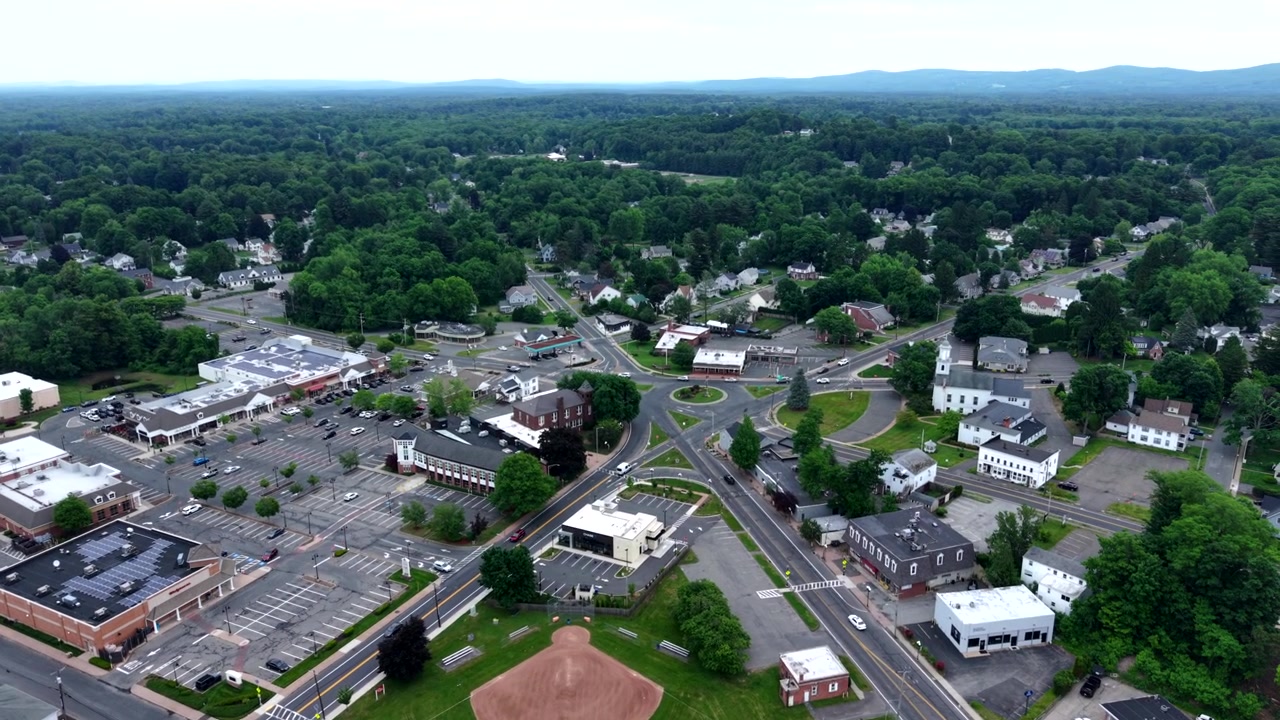 Drone kredser til højre over East Longmeadow Rådhus rundkørsel, der afslører et frodigt grønt forstadssamfund med boliggader, handelsområde og fjern bjergkæde i Massachusetts