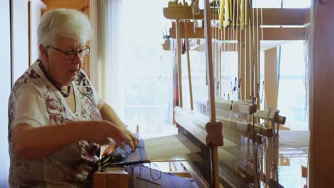 Side-view-of-old-caucasian-senior-woman-weaving-cloth-on-handloom-machine-in-a-workshop-4k