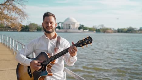 Adult-man-and-musician,-singer,-guitarist,-and-artist-is-standing-at-a-park-singing-a-song-and-strumming-his-guitar-with-the-thomas-jefferson-memorial-in-the-background