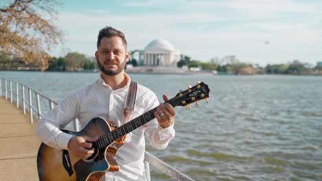 Hombre-Adulto-Y-Músico,-Cantante,-Guitarrista-Y-Artista-Está-Parado-En-Un-Parque-Cantando-Una-Canción-Y-Tocando-Su-Guitarra-Con-El-Memorial-De-Thomas-Jefferson-En-El-Fondo