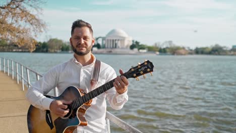 hombre adulto y músico, cantante, guitarrista y artista está de pie en un parque cantando una canción y tocando su guitarra con el monumento a thomas jefferson en el fondo