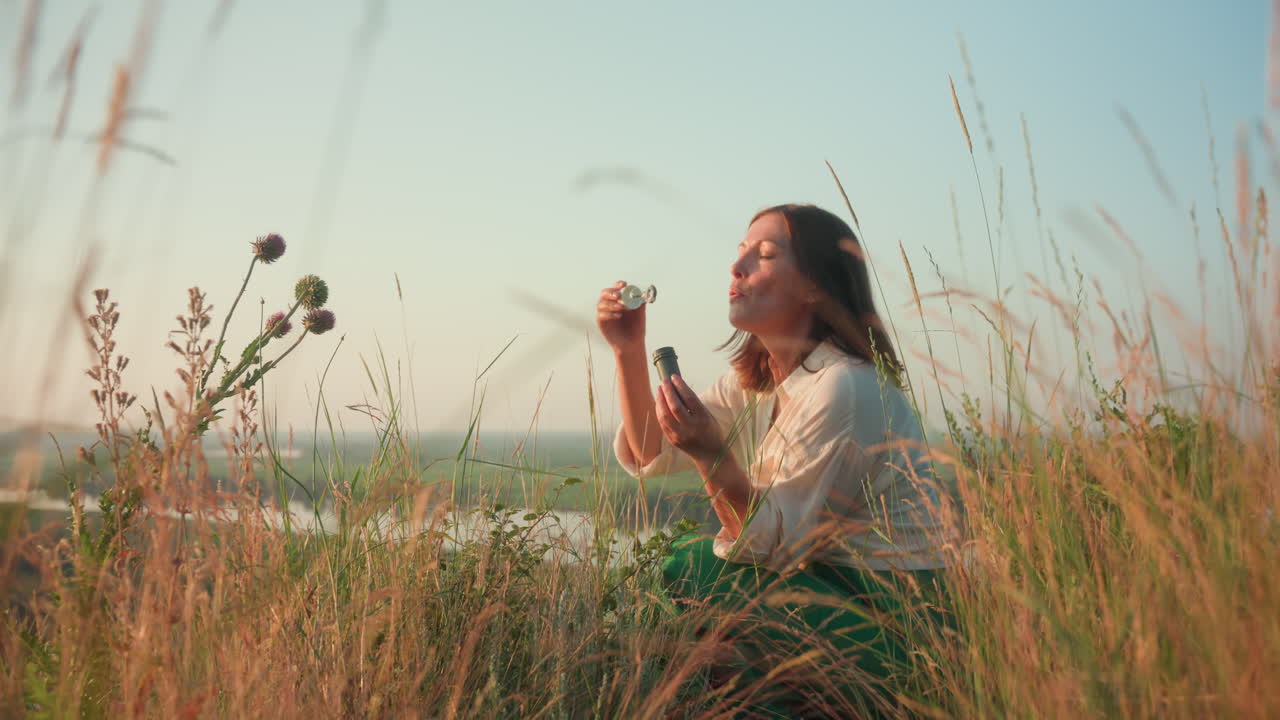 Sunset lit woman in green skirt squatting on grassy river bank opening bubble bottle then blowing shimmering soap bubbles that float over blurred valley and wildflowers